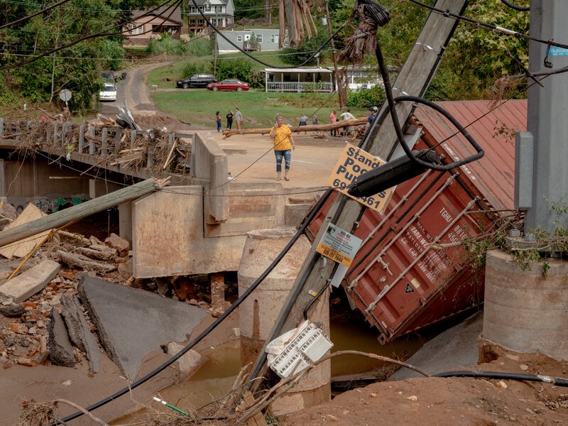 Flood-damaged bridge with collapsed sections and a derailed railcar in muddy water, as residents survey storm destruction in a nearby neighborhood.