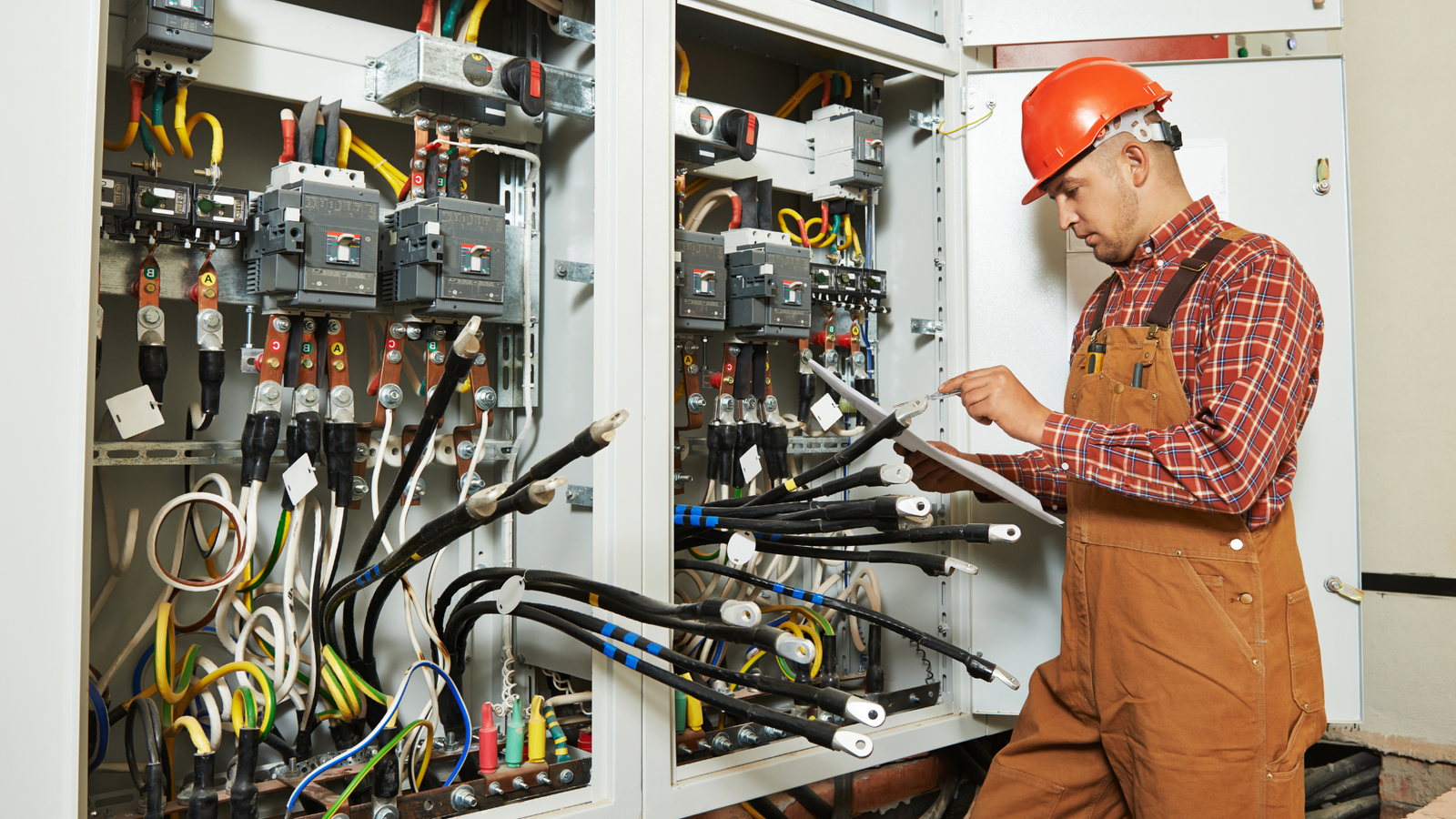 Electrician in hard hat inspecting industrial electrical panel with wiring and circuit breakers while reviewing checklist on clipboard.