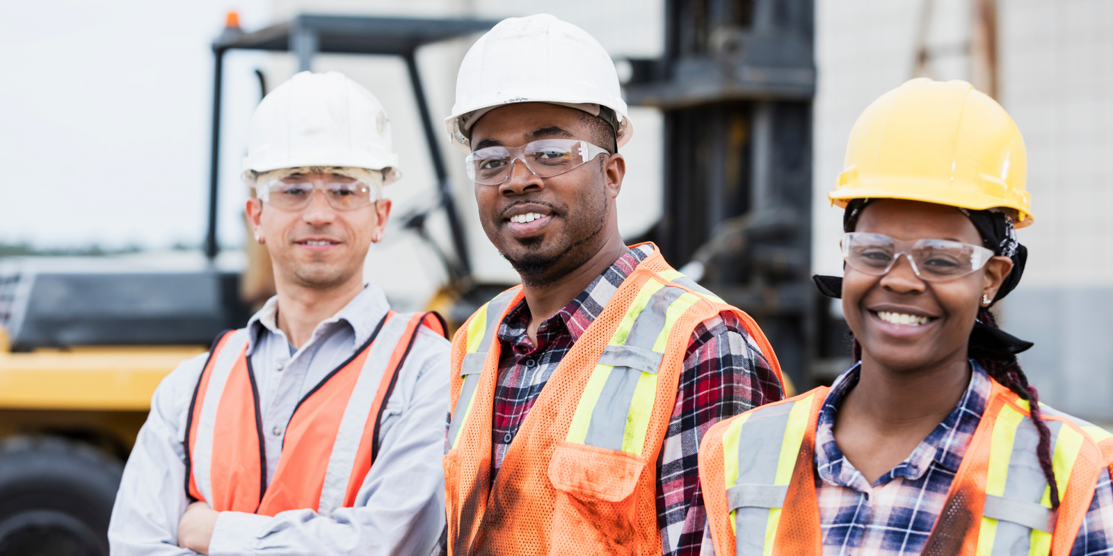 Three construction workers wearing hard hats, safety glasses, and high-visibility vests standing together at a job site with heavy equipment in the background